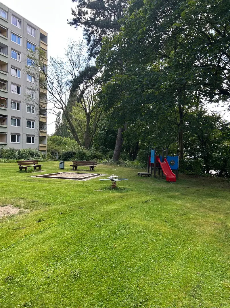 Spielplatz mit Sandkasten, Bänken und roter Rutsche auf einer gemähten Rasenfläche, links ein Wohnhochhaus, ringsum hohe Bäume
