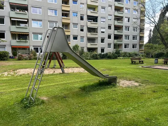 Metallrutsche auf einer Sandfläche vor einem Wohnhochhaus mit Balkonen, der Rasen ringsum gemäht